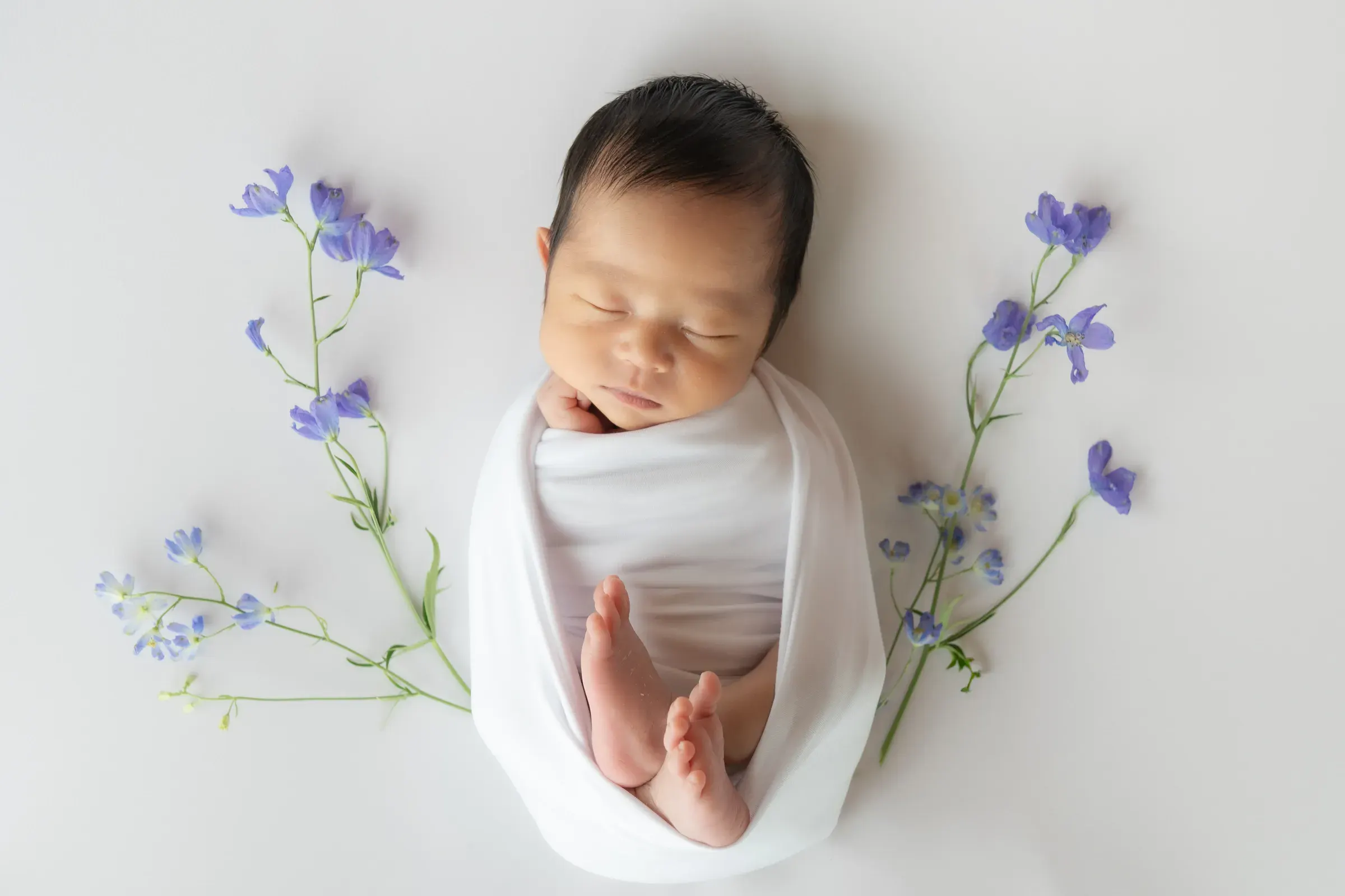 白いおくるみに包まれ、紫の小花に囲まれて眠る赤ちゃん