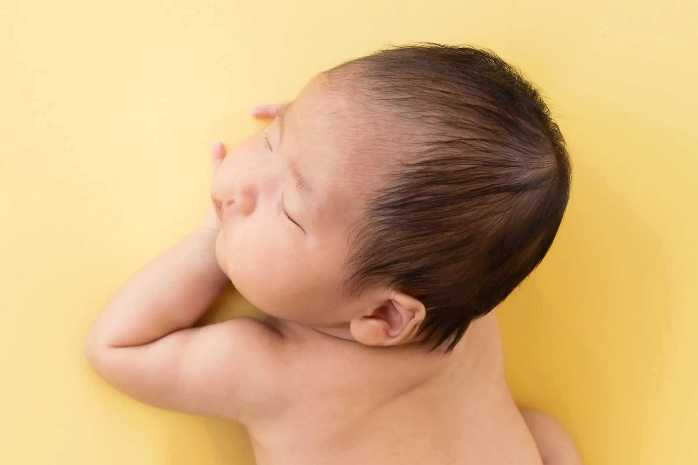 黄色い背景の上で、腕に顔をのせて眠る赤ちゃんの横顔